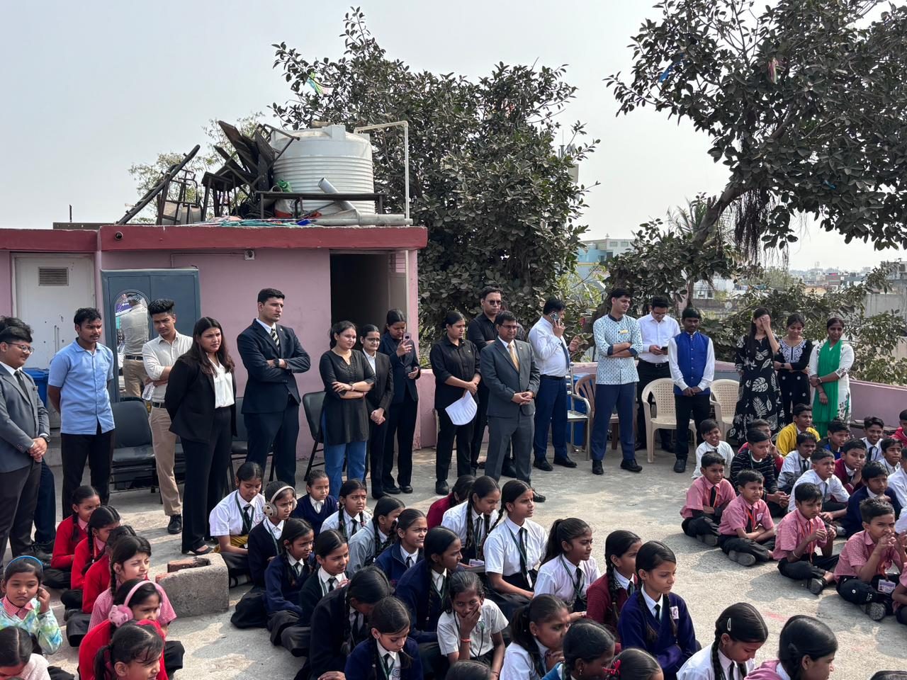 Students seated on a school terrace for an assembly, with visiting staff standing alongside them.